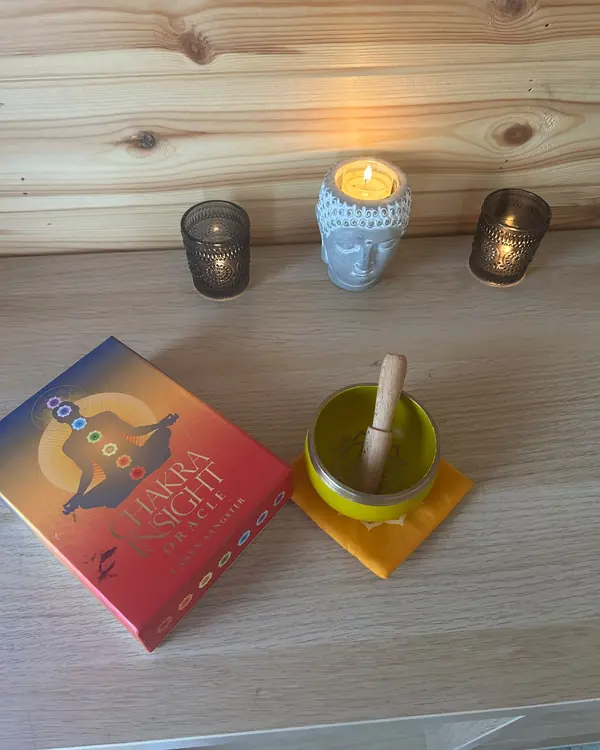 A candle in a Buddha holder, a chakra oracle card deck and a singing bowl on a wooden shelf